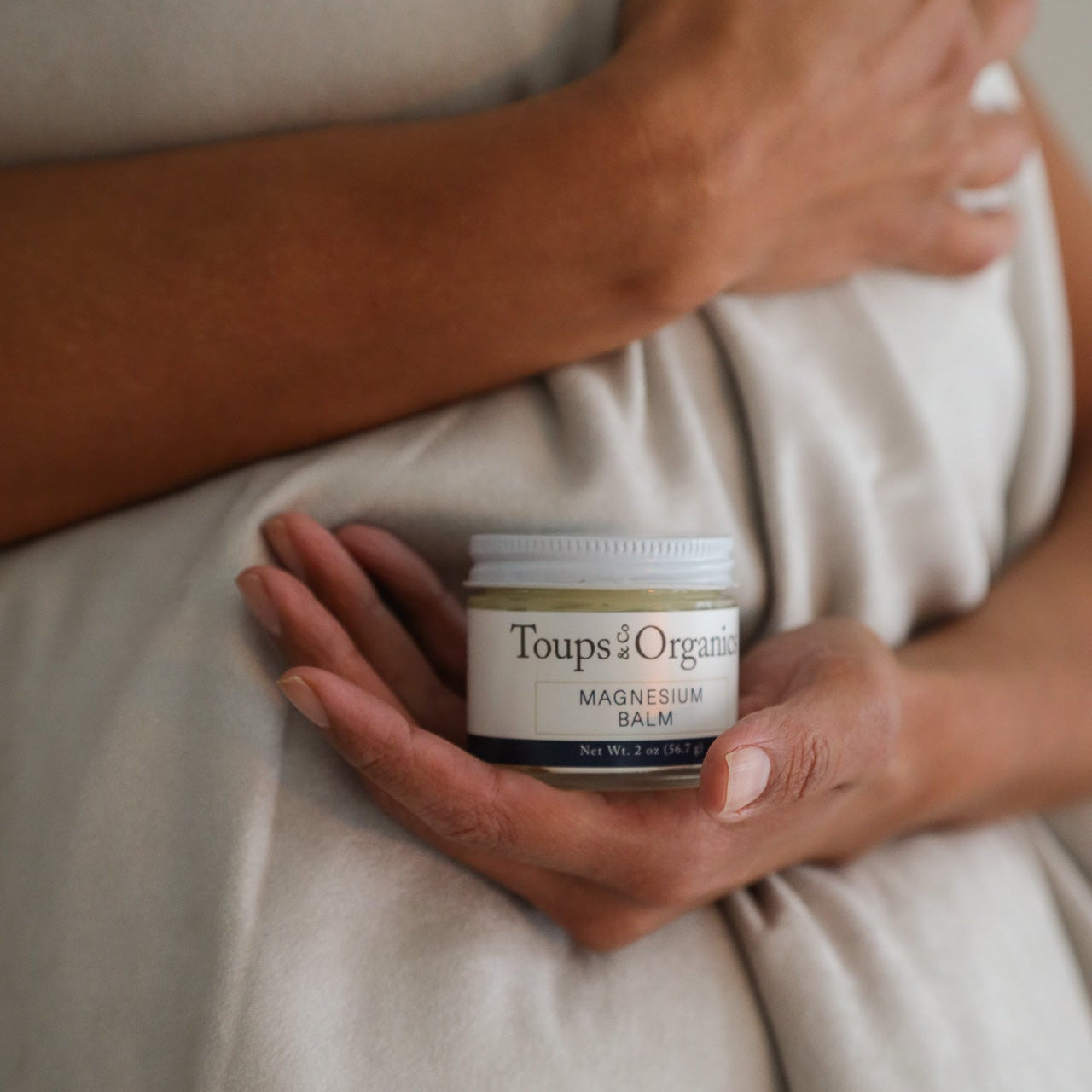 Someone holds a jar of Toups and Co Organics Magnesium Balm in front of a beige fabric background.