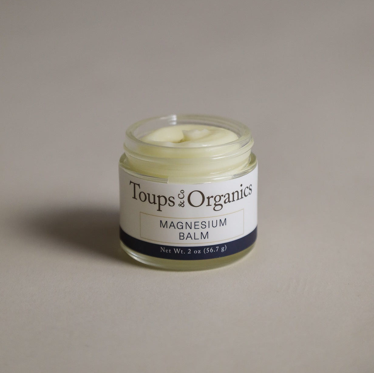 A small glass jar of Toups and Co Organics Magnesium Balm containing white cream sits on a plain background, with a label displaying the product name and net weight.