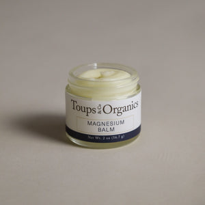 A small glass jar of Toups and Co Organics Magnesium Balm containing white cream sits on a plain background, with a label displaying the product name and net weight.