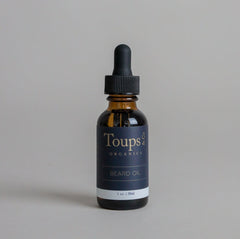 A brown glass bottle of Toups and Co Organics Beard Oil, formulated to promote beard growth and moisturize facial hair, is set against a simple gray background. The black dropper cap provides an elegant accent to this must-have grooming product.