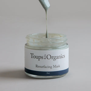 A jar labeled Toups and Co Organics Resurfacing Mask, with a spatula lifting light green cream from the open container, enriched with blue algae extract for powerful anti-aging benefits.