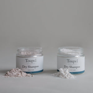 Two jars of Toups and Co Organics Dry Shampoo, one labeled Dark Hair and the other Light Hair, are placed side by side with small mounds of powder in front of each jar on a simple background, showcasing their effective oil-absorbing properties for healthy scalp care.