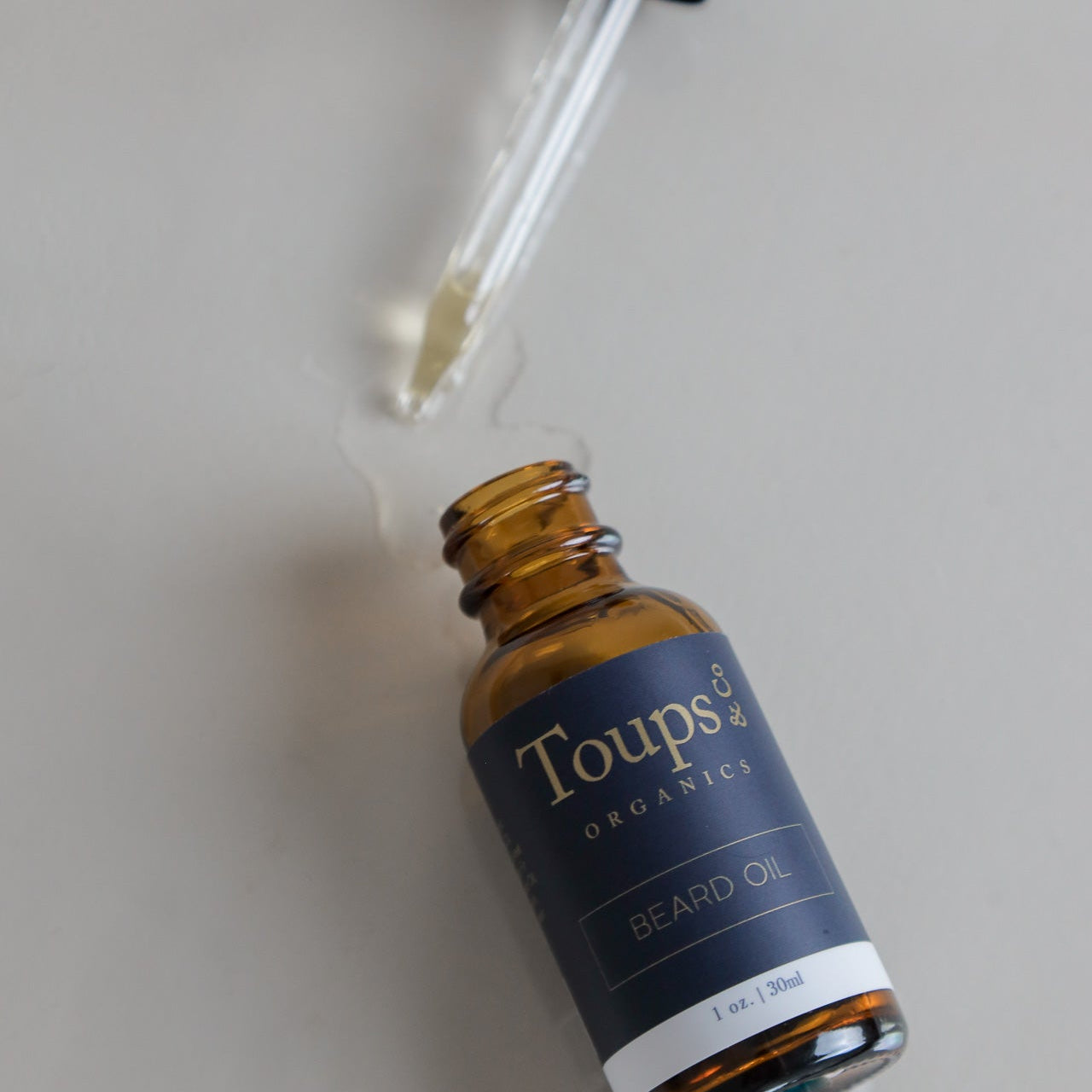 A brown glass bottle of Toups and Co Organics Beard Oil sits open on a surface, its dropper resting beside it and a small pool of oil spilled nearby, showcasing this nourishing beard oil—an essential moisturizer crafted to support healthy beard growth.