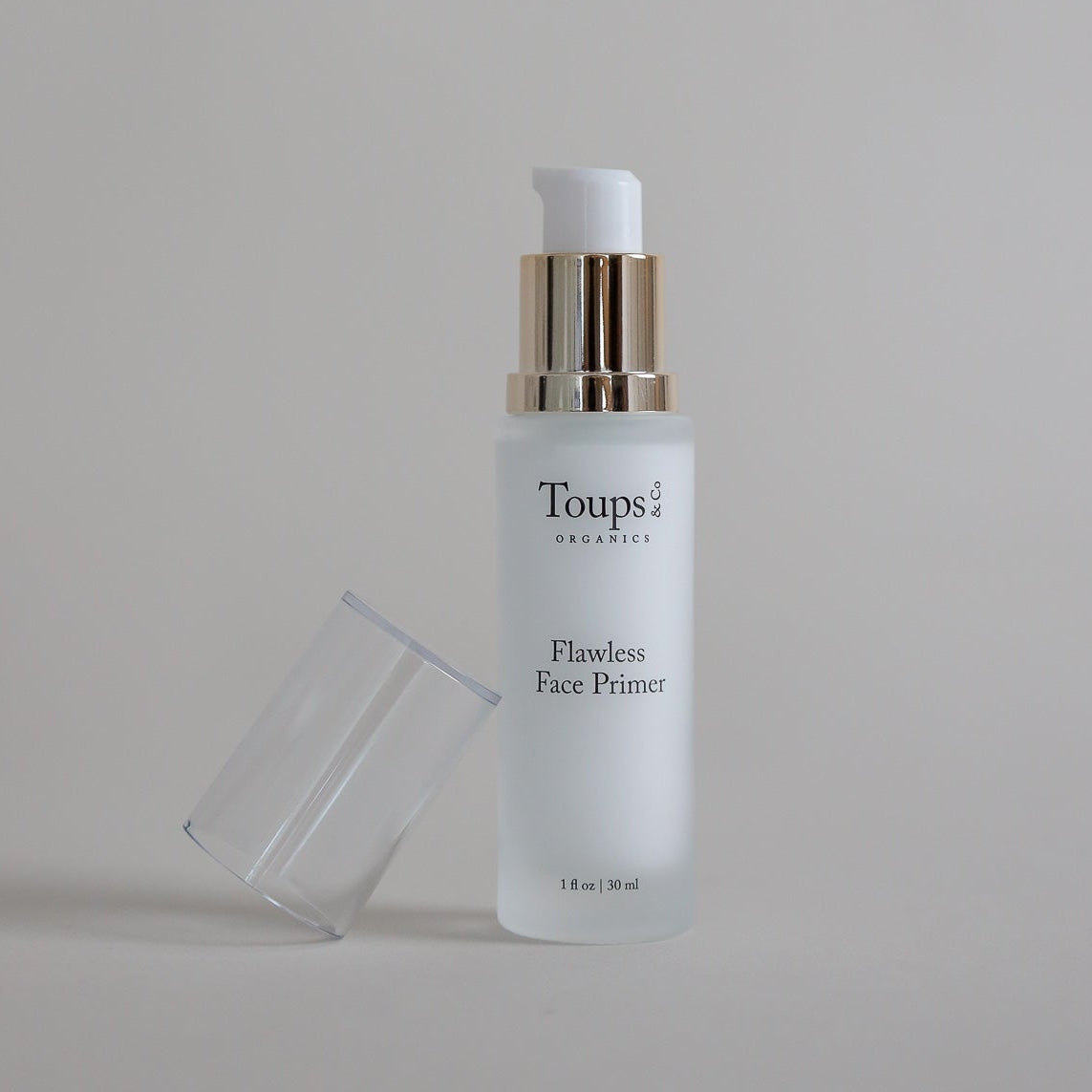 A bottle of Toups and Co Organics Flawless Face Primer with a pump dispenser rests on a neutral surface with the top off.
