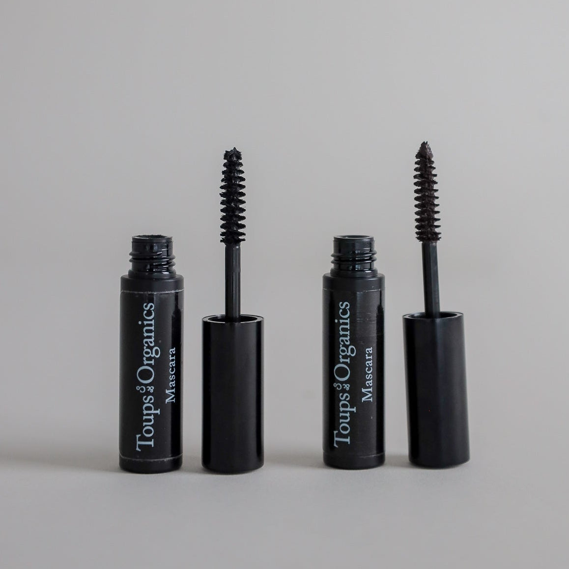 Product photography featuring two open mascara tubes with applicator brushes removed and displayed on a clean grey seamless background, highlighting the makeup product and mascara wands.