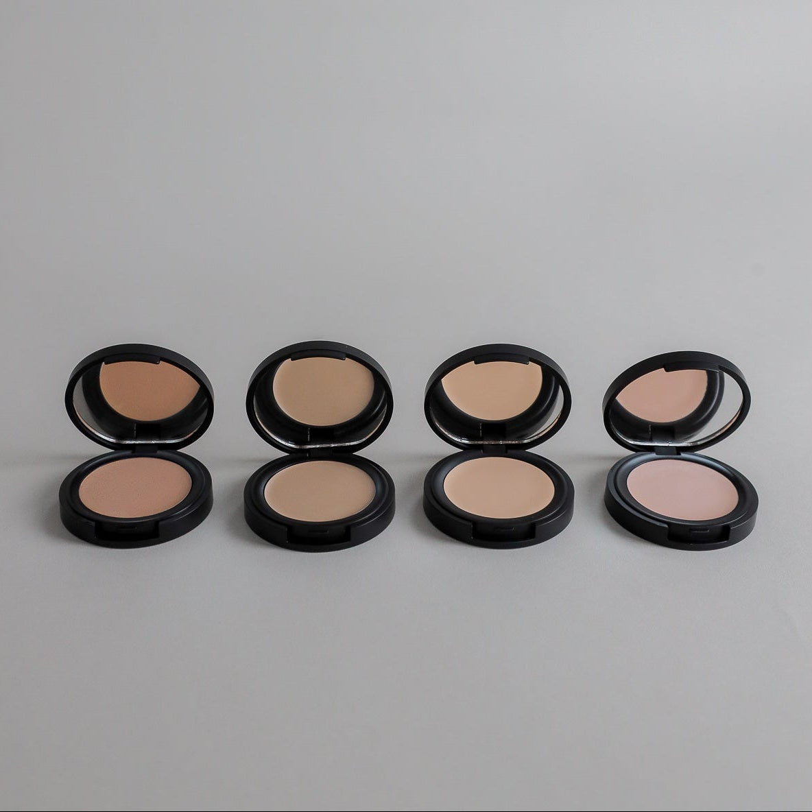 Four Toups & Co non-toxic concealers displayed open to showcase a range of complexion shades against a clean grey seamless background. A simple, modern product photo highlighting natural makeup and versatile beauty essentials.