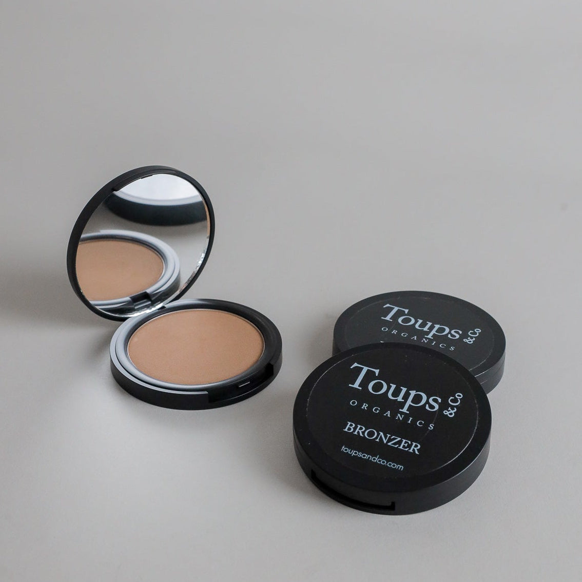Toups & Co bronzer compacts photographed open and closed on a clean grey seamless background, showcasing the bronzer formula and classic black packaging. A minimalist beauty product image highlighting natural-looking complexion makeup.
