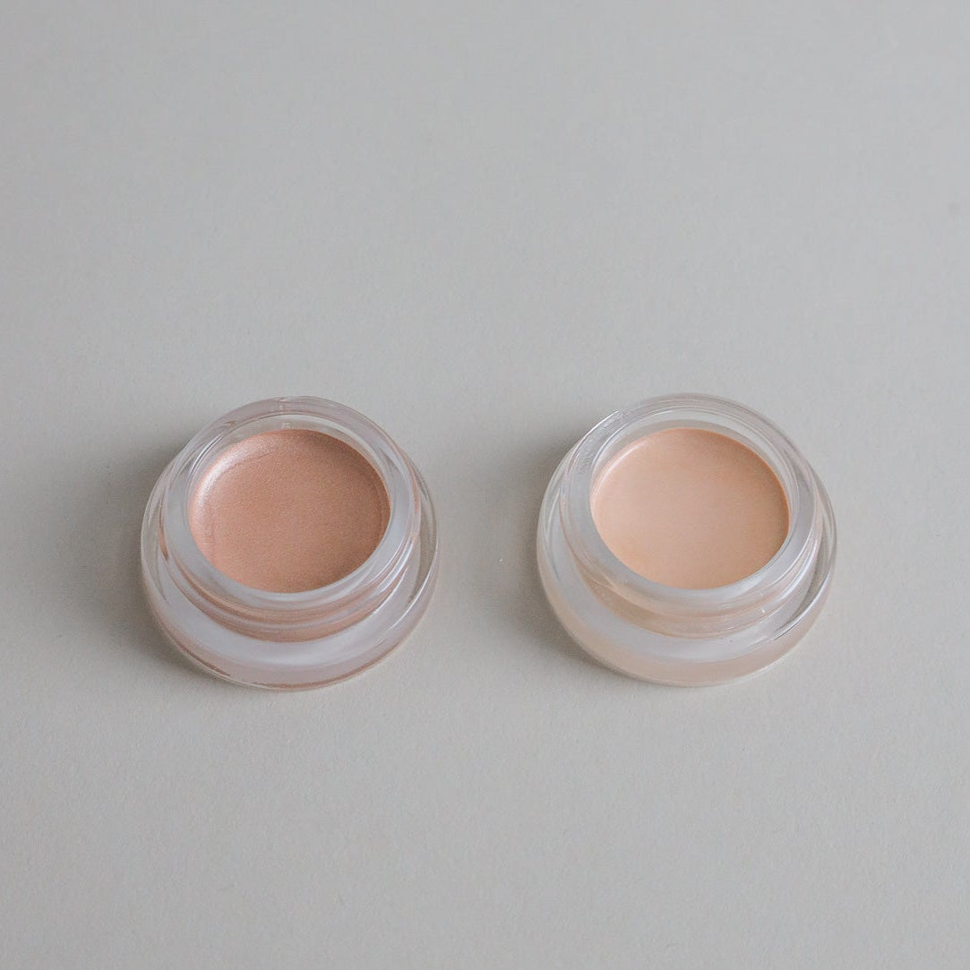 Two small open Cream Luminizer Pots by Toups and Co Organics, labeled Champagne (right) and Rose Gold (left), sit on a tan surface with black lids beside them—made with non-toxic ingredients for a natural-looking glow.