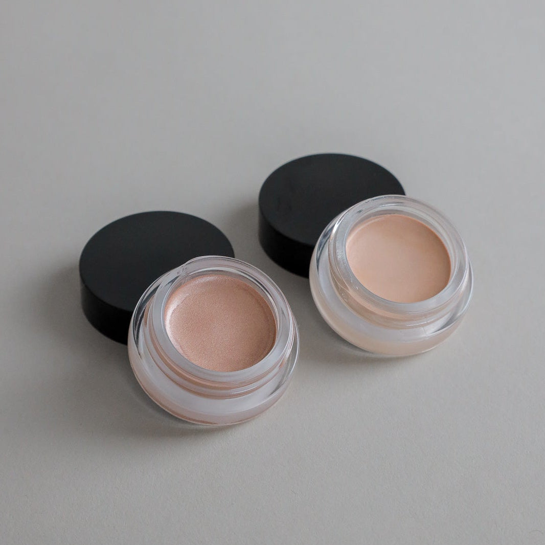 Two small open Cream Luminizer Pots by Toups and Co Organics, labeled Champagne (right) and Rose Gold (left), sit on a tan surface with black lids beside them—made with non-toxic ingredients for a natural-looking glow.