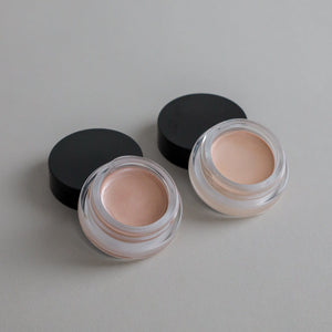 Two small open Cream Luminizer Pots by Toups and Co Organics, labeled Champagne (right) and Rose Gold (left), sit on a tan surface with black lids beside them—made with non-toxic ingredients for a natural-looking glow.