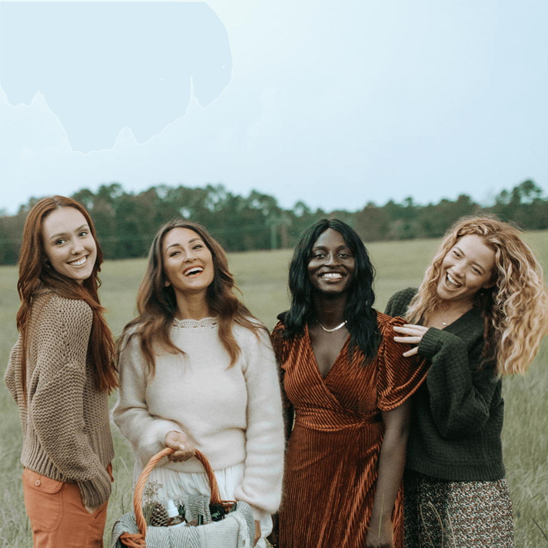 Four women smiling together in a field, with one holding a picnic basket.