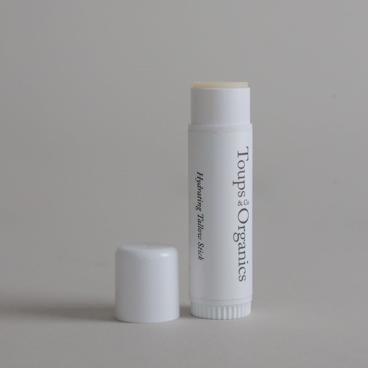 A white tube labeled Hydrating Tallow Stick from Toups and Co Organics, made with nourishing grass-fed tallow, stands upright with its cap placed nearby on a plain light gray background.