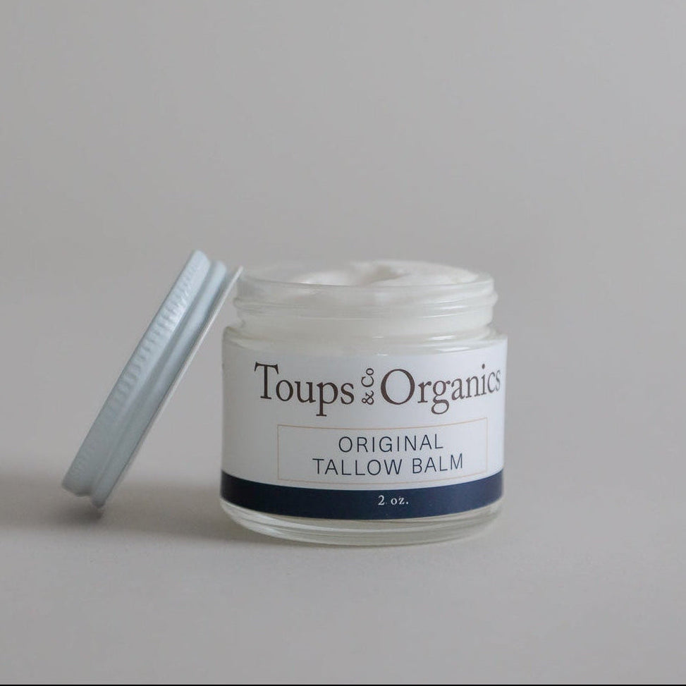 A glass jar of Toups and Co Organics Tallow Balm - Original, 2 oz, with the lid off, shows rich moisture for dry sensitive skin on a plain light background.
