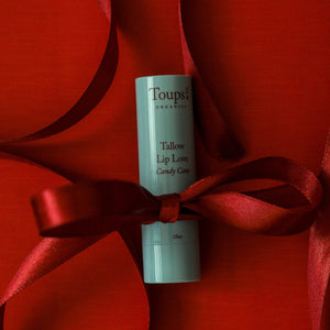A tube of Toups and Co Organics Candy Cane Lip Love, made with nourishing grass-fed tallow, stands upright with a red satin ribbon on a red background.