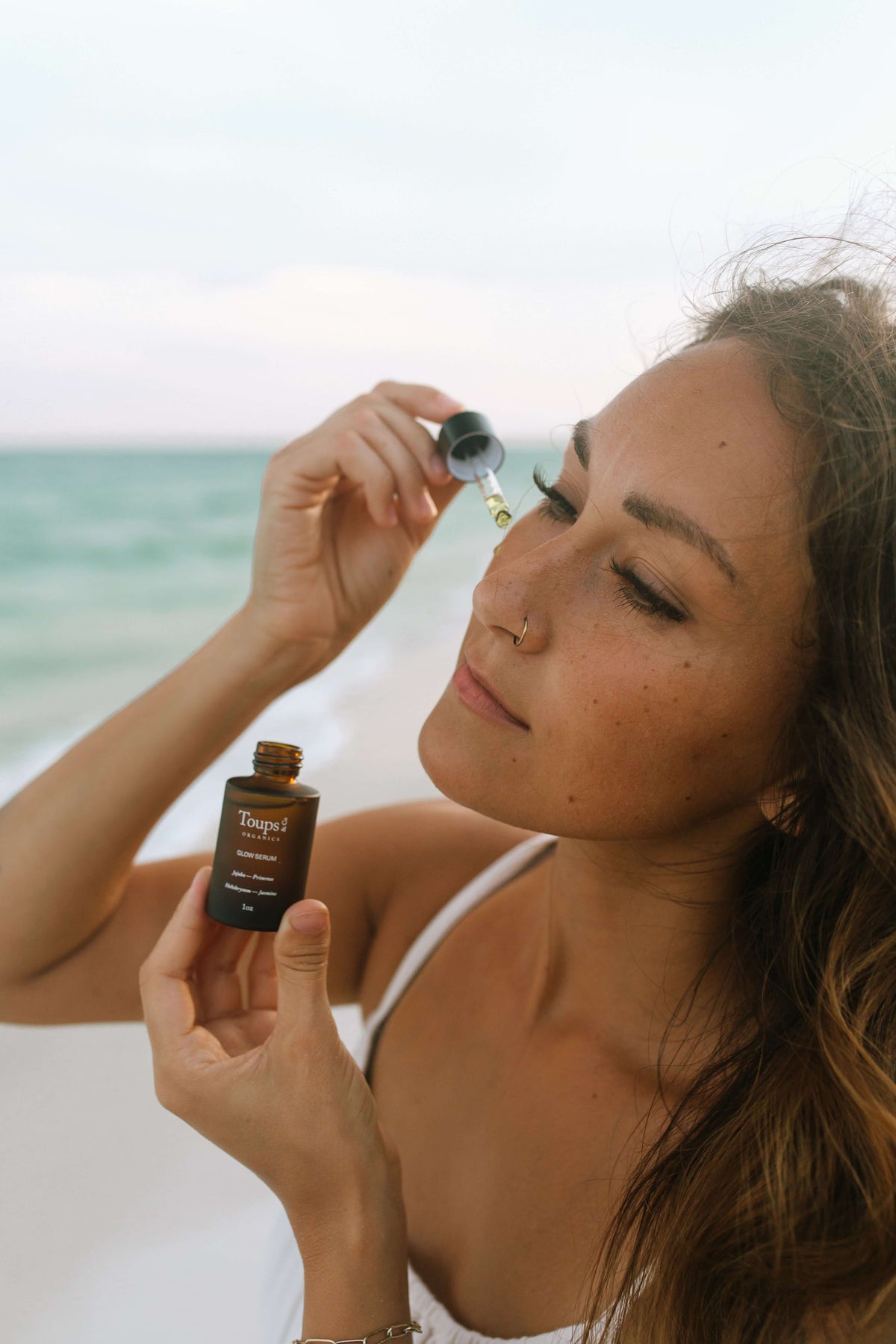 A woman holding a bottle of Toups Organics Glow Serum and applying a drop to her cheek on a beach.