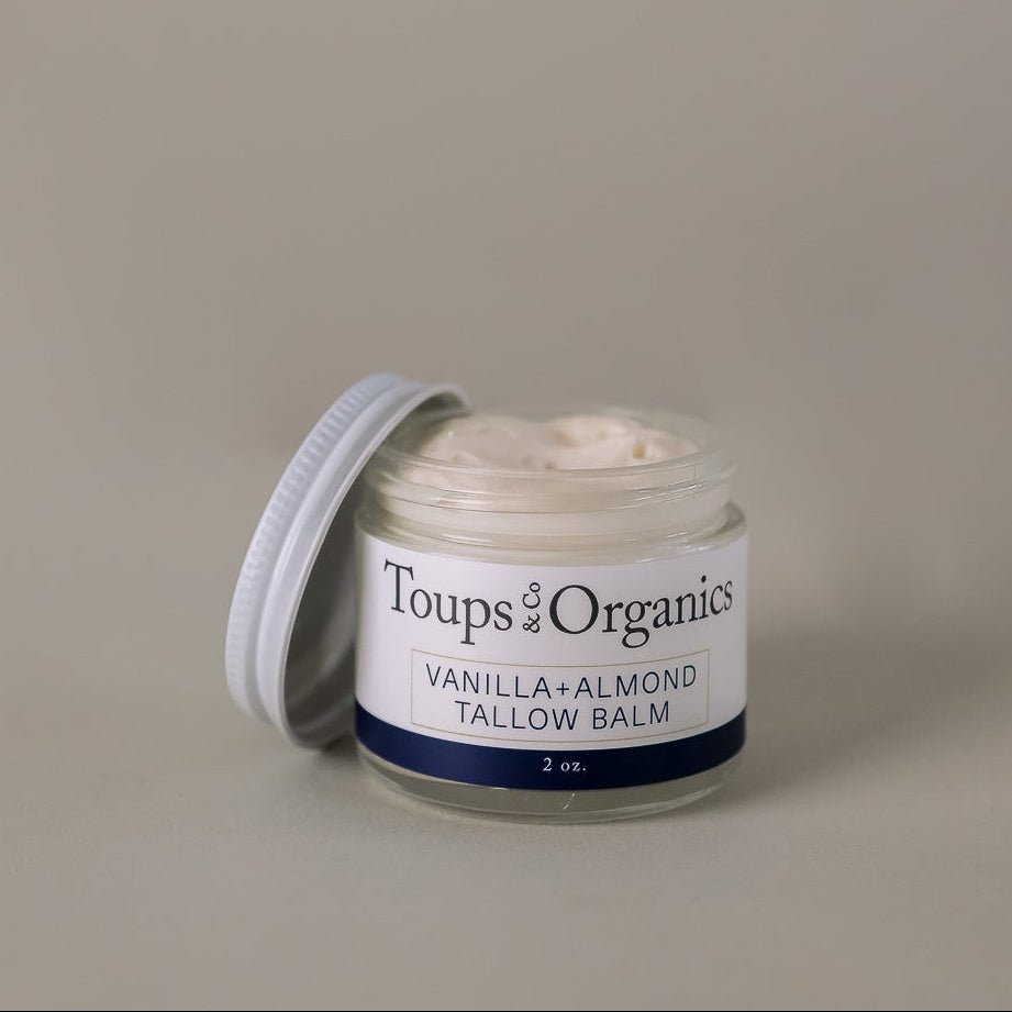 A glass jar of Toups and Co Organics Tallow Balm - Vanilla + Almond, made with nourishing grass-fed tallow, sits with the lid off to reveal the creamy balm inside, inviting you to experience ultimate skin hydration against a plain background.