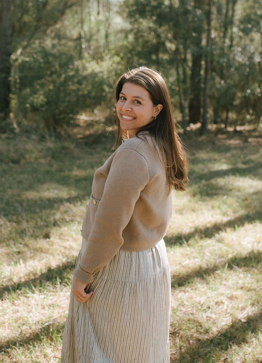 A woman in a beige cardigan and long skirt standing in a wooded area, smiling over her shoulder at the camera.